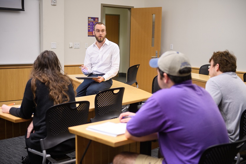 Professor leans on a desk while lecturing a small class